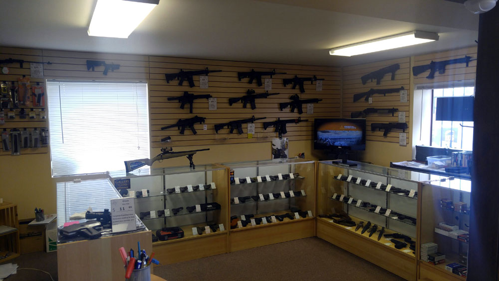 Single Category 3 A gun store interior with rifles mounted on the walls, handguns displayed in glass cases, and accessories on shelves. Fluorescent lights illuminate the room and a window lets in natural light.
