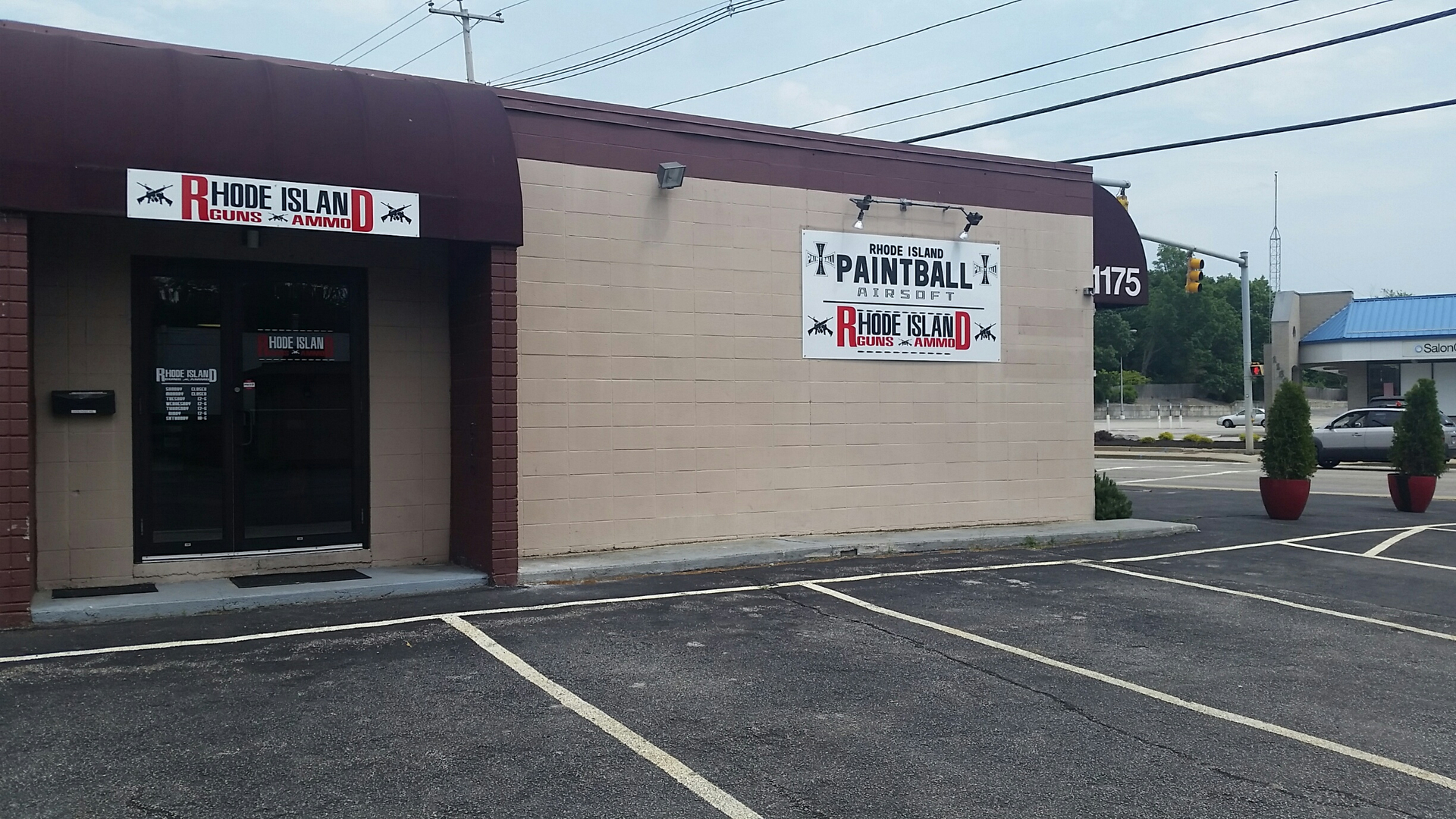 A tan brick building with signs reading "Rhode Island Guns & Ammo" and "Rhode Island Paintball & Airsoft." The entrance is on the left, and there are empty parking spaces and potted shrubs in front.