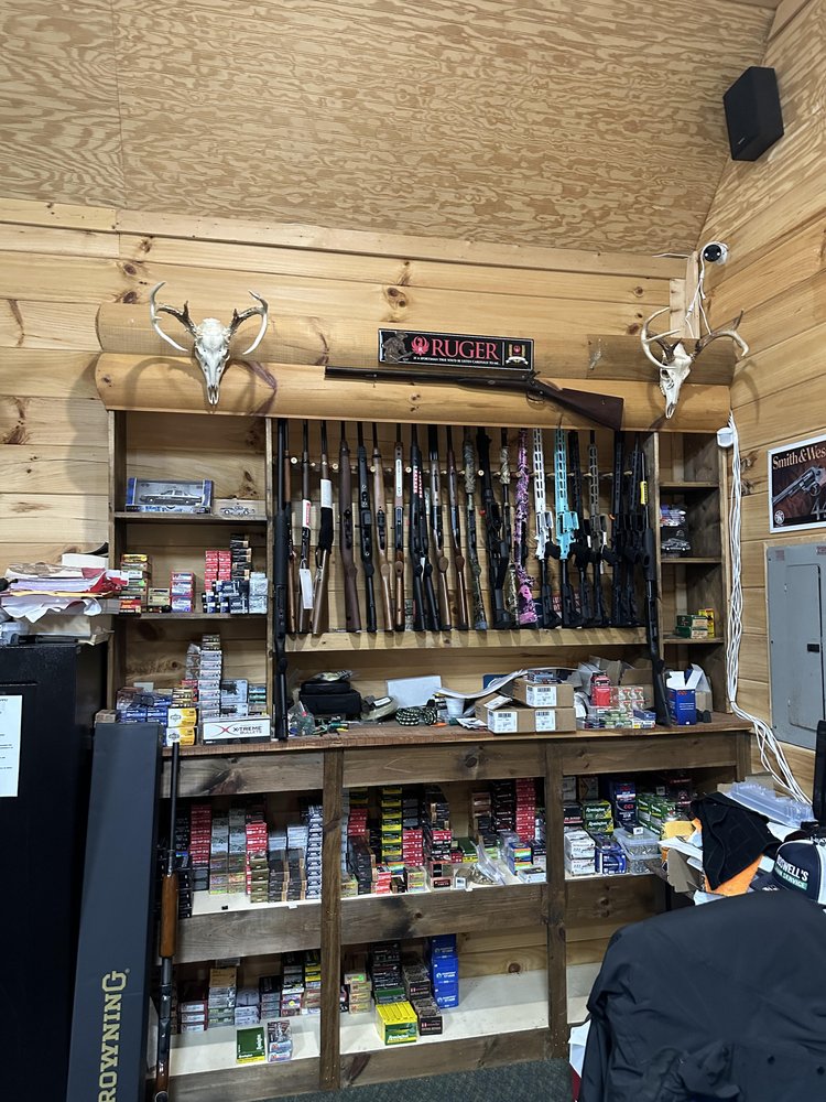 A wooden wall display holds several rifles and shotguns, flanked by two mounted deer skulls with antlers. Below are shelves filled with various boxes of ammunition and other firearm accessories.