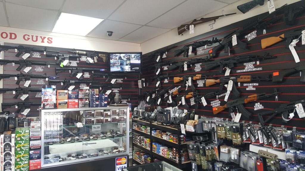 A gun store interior with rifles and guns displayed on wall racks, ammunition and accessories on shelves, a glass case with handguns, and a security camera monitor above.