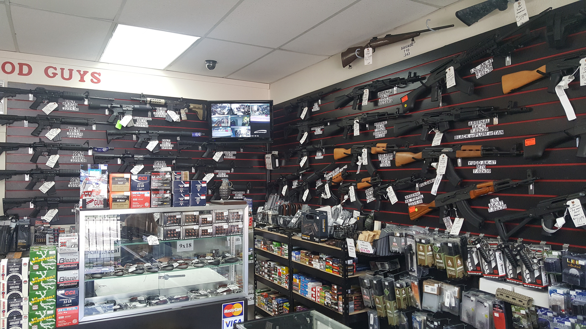 A gun store interior with rifles and guns displayed on wall racks, ammunition and accessories on shelves, a glass case with handguns, and a security camera monitor above.