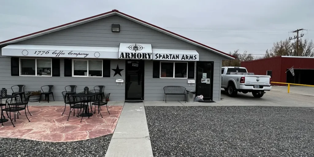 A gray building with a sign reading "ARMORY SPARTAN ARMS" and "1776 Caffe Company." Patio tables and chairs are outside, with a white pickup truck parked to the right and a red building in the background.