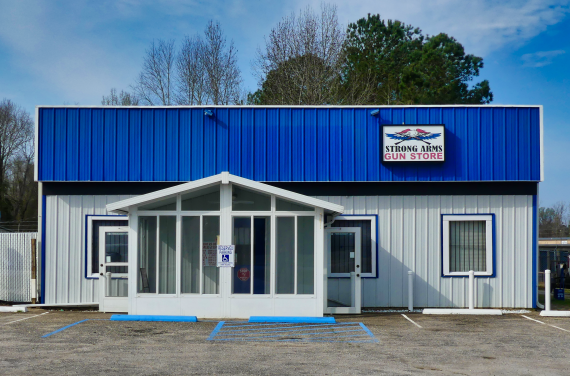 Single Category 1 A small, blue-and-white building with a sign reading "Strong Arms Gun Store" above the entrance, flanked by trees and a clear sky, with two accessible parking spaces in front.
