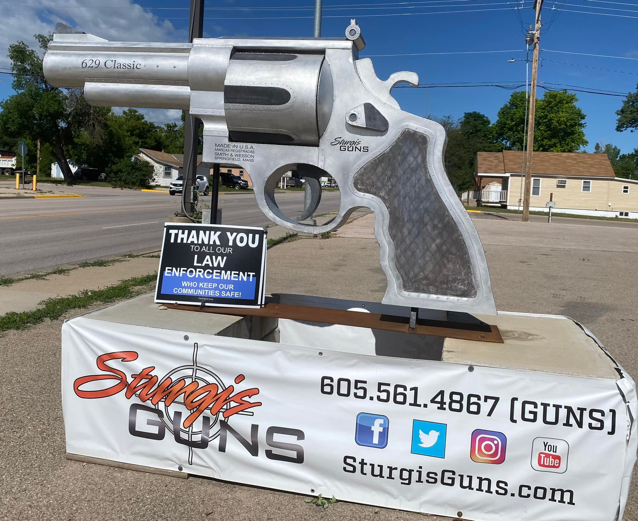 Single Category 5 A large metal sculpture of a revolver is displayed on a street corner. A sign thanking law enforcement is in front, and a banner below advertises "Sturgis Guns" with contact info and social media icons.
