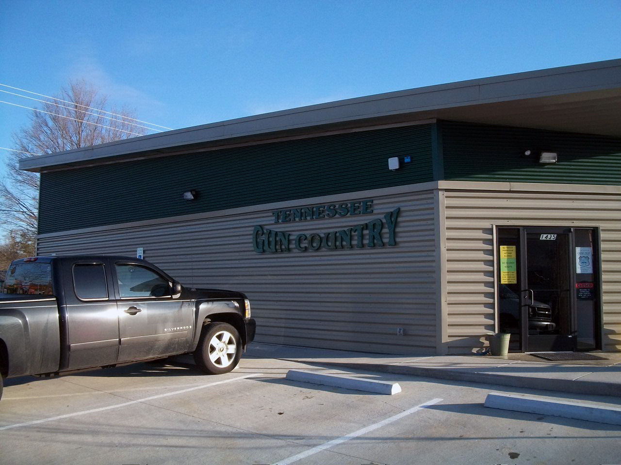 Firearms Training Directory 11 A black pickup truck is parked outside a single-story building with a sign reading "Tennessee Gin Company." The building has green and beige siding, large windows, and a clear blue sky above.