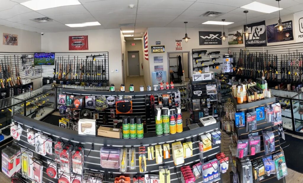 A gun store interior with rifles and firearms displayed on wall racks, shelves stocked with ammunition, accessories, and cleaning supplies, and glass display cases under bright fluorescent lighting.
