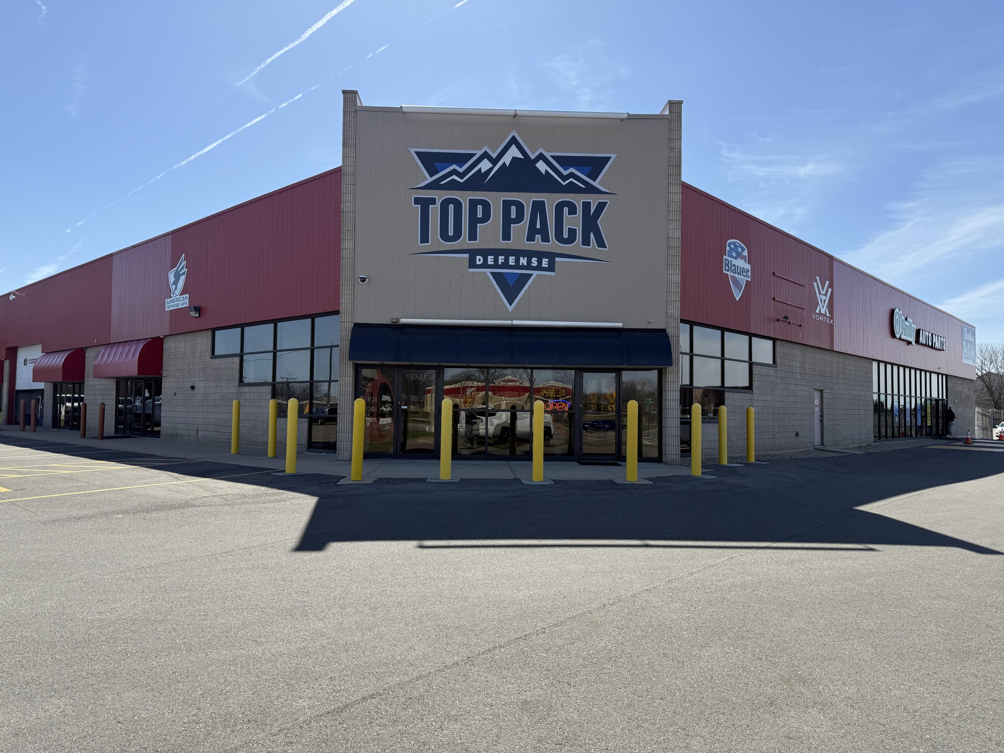 Shooting Range Directory 28 A large commercial building with a sign reading "Top Pack Defense" on the front facade. The building has red accents, glass doors, and yellow bollards at the entrance. The sky is clear and blue.