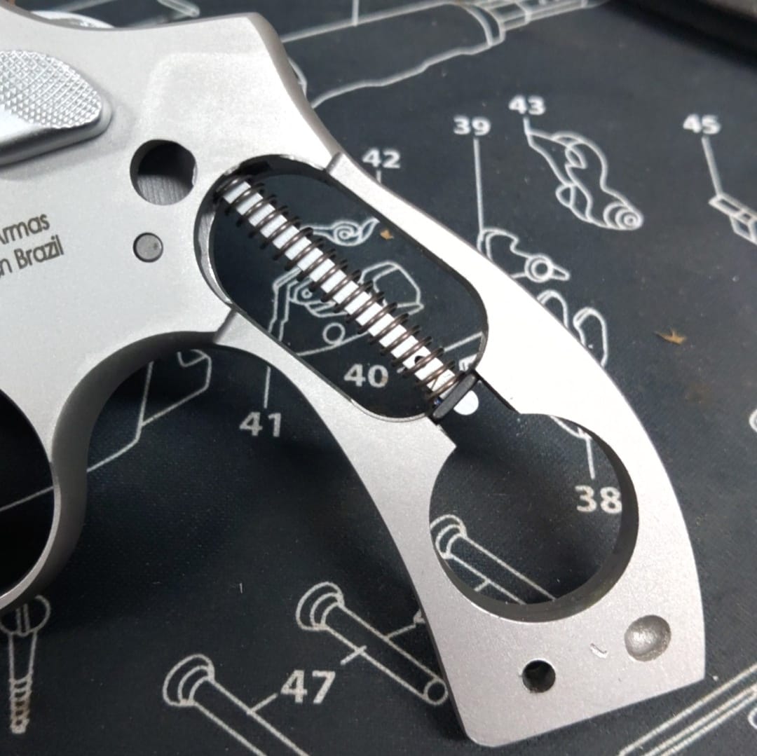 Close-up of a revolver frame showing the internal spring mechanism, placed on a black gun maintenance mat with white technical diagrams and numbered gun parts.