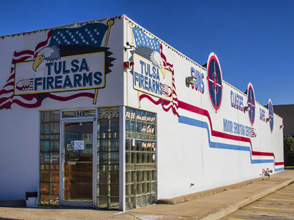 Oklahoma Firearms Training 4 A white building with patriotic murals, including American flags and an eagle, displays signs reading "Tulsa Firearms" and features target symbols on the exterior wall. The entrance has glass blocks and a door with signage.