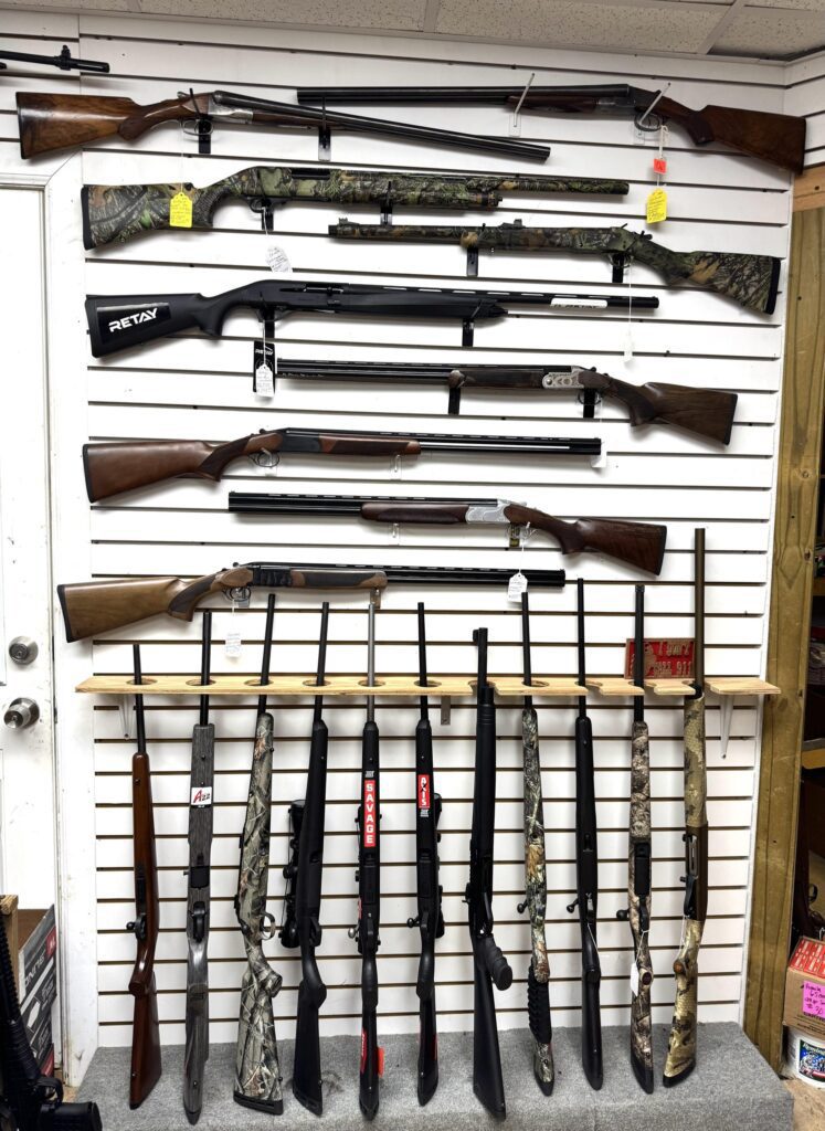 Several rifles and shotguns are displayed on a white slat wall, with more firearms standing upright on a rack below. Some firearms have camouflage patterns; price tags are attached to a few. Boxes of ammo are visible on the shelf.