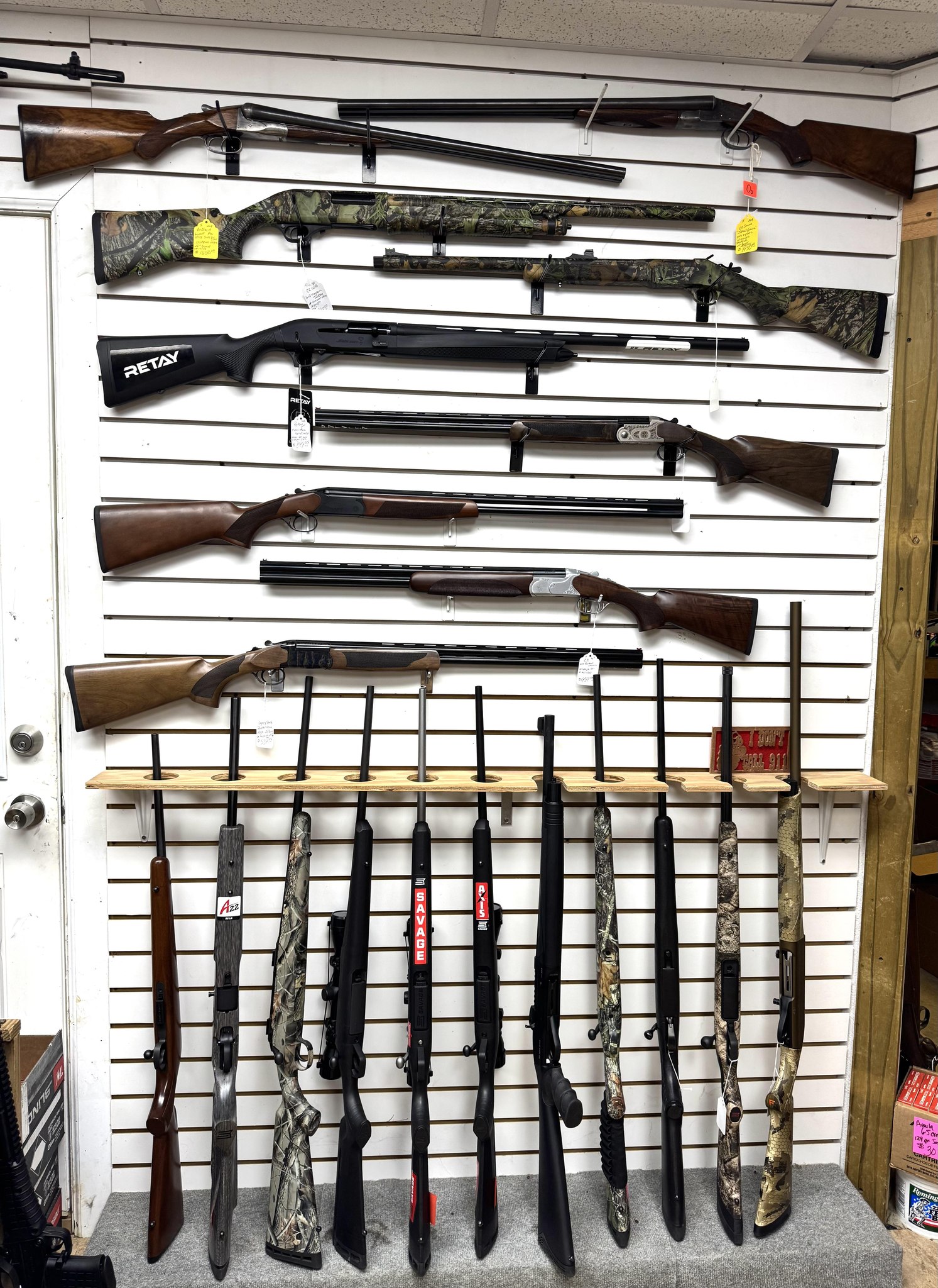 Single Category 1 Several rifles and shotguns are displayed on a white slat wall, with more firearms standing upright on a rack below. Some firearms have camouflage patterns; price tags are attached to a few. Boxes of ammo are visible on the shelf.