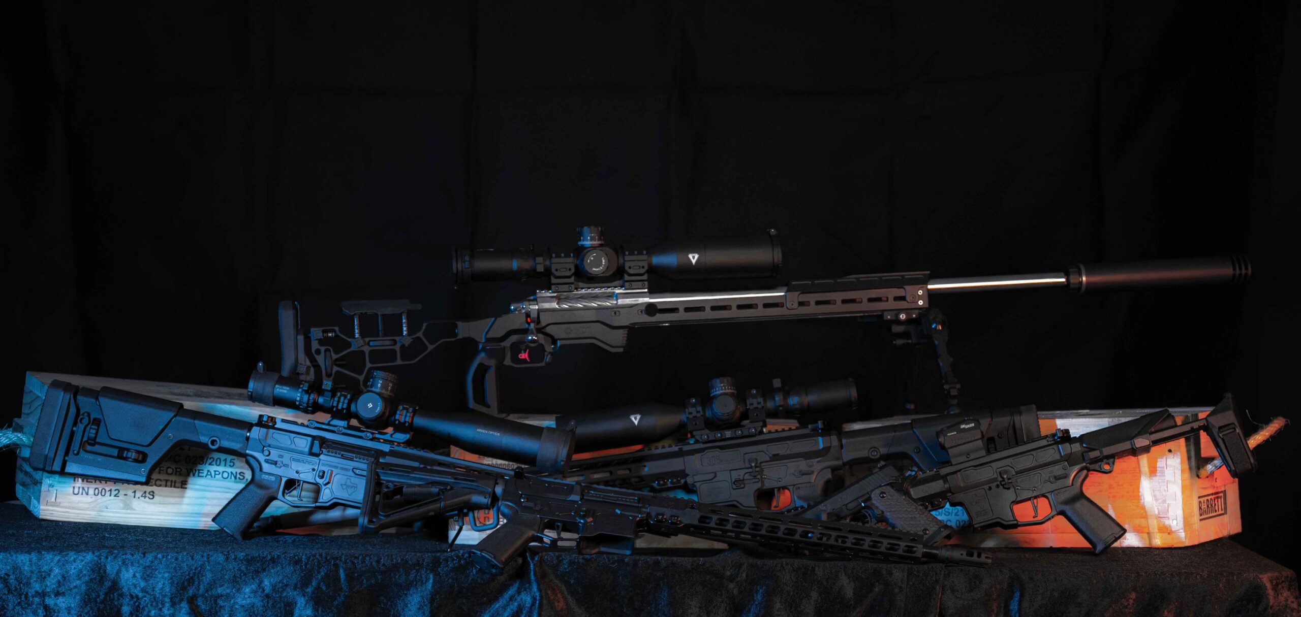 Single Category 5 Three modern rifles with scopes and tactical accessories are displayed on a black surface, with a wooden crate beneath them, all set against a dark background with blue and orange lighting accents.