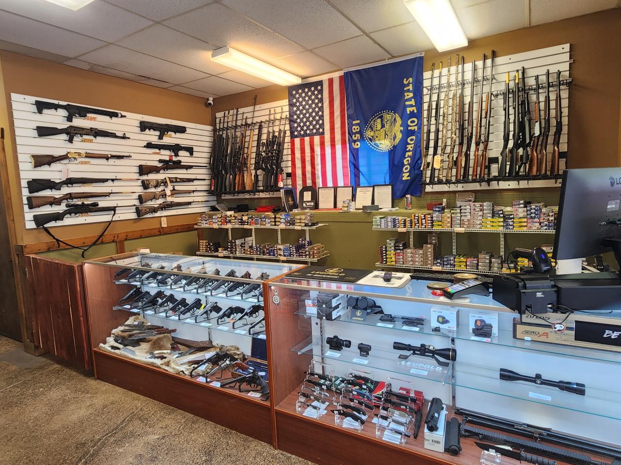Oregon Firearms Training 4 A gun shop interior with rifles and shotguns displayed on the wall, handguns in glass cases, various ammunition boxes on shelves, and an American flag and Oregon state flag hanging behind the counter.