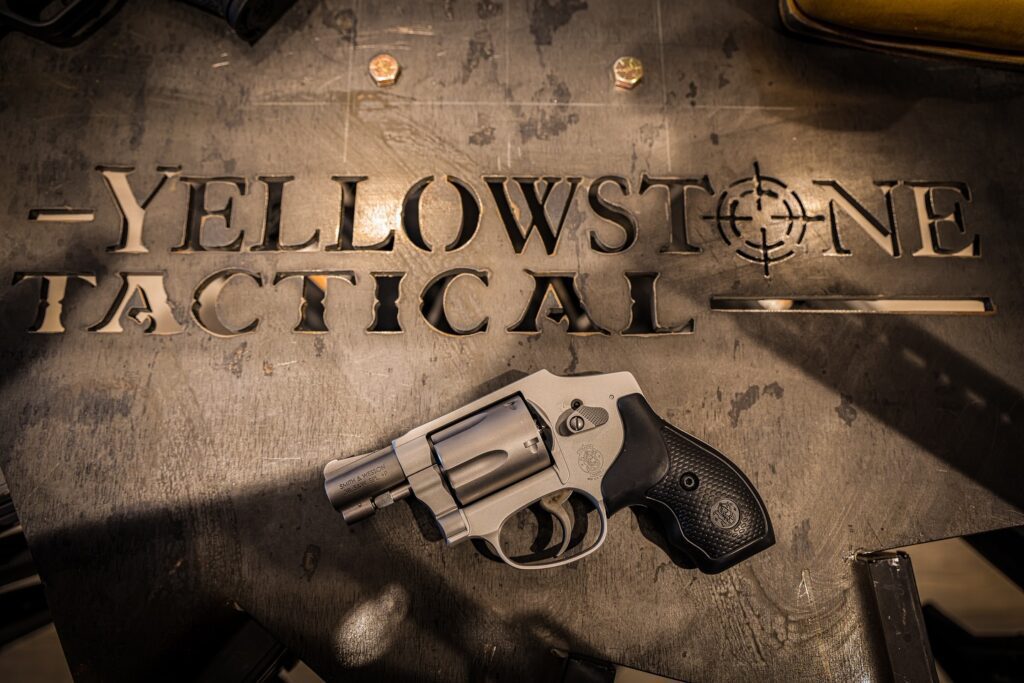 A silver and black revolver rests on a metal surface cut with the words "Yellowstone Tactical" and a target symbol. Two bolts are also visible on the table.