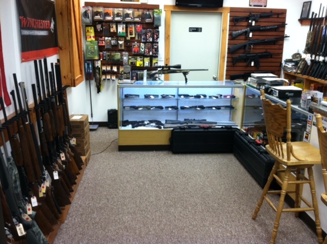 A gun shop interior with rifles displayed on racks, firearms in a glass display case, accessories on shelves, and a wooden chair in front of the counter.