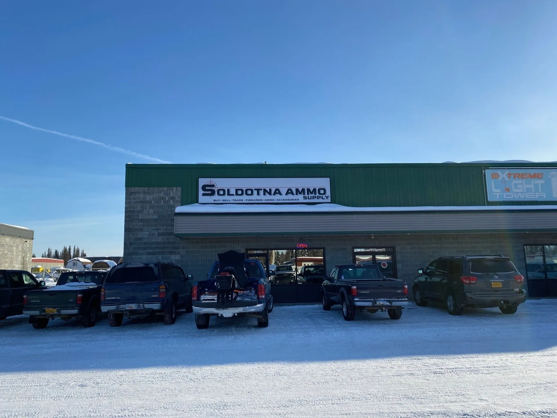 Ammunition supply store with a sign reading "Soldotna Ammo Supply" above the entrance. Several vehicles are parked in front on a snowy lot under a clear blue sky.
