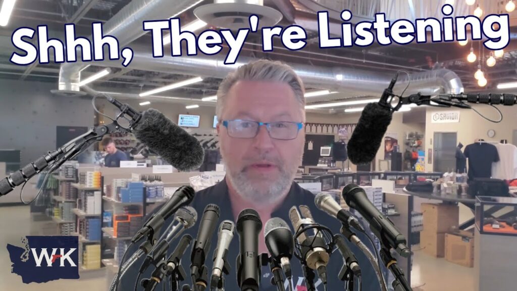 A man with glasses stands in an office, surrounded by microphones. Large text above him reads, "Shhh, They're Listening." The background shows desks, shelves, and office equipment.