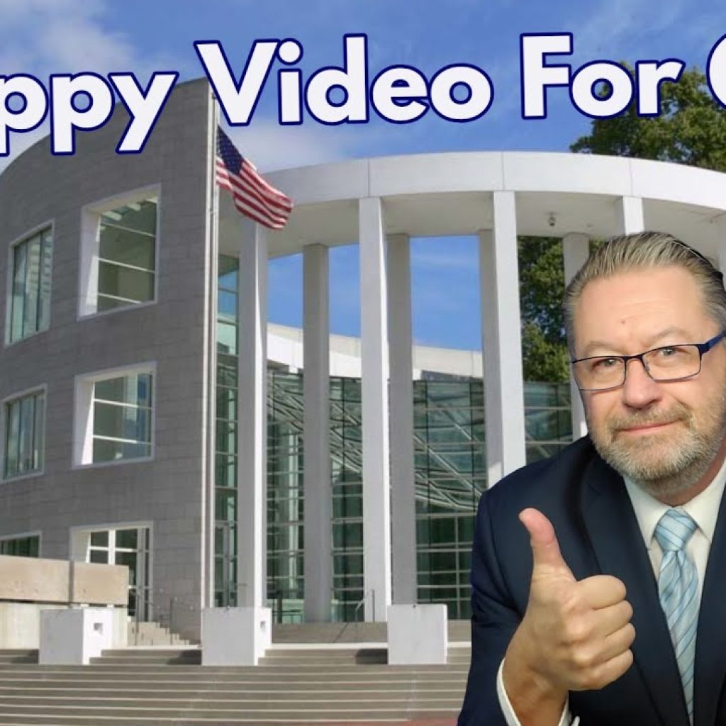 A man in a suit and glasses gives two thumbs up in front of a modern courthouse. The text reads "A Happy Video For Once." An American flag is visible. The lower left has a logo with "WK.