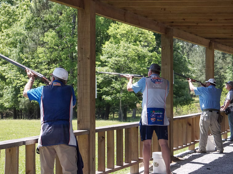 Alabama Firearms Training 6 Three people at an outdoor shooting range stand under a covered area, aiming shotguns at targets. Trees and greenery are visible in the background on a sunny day.