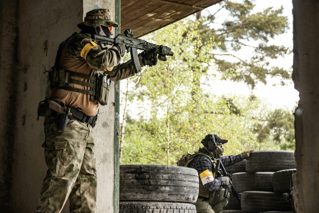 Two armed soldiers in camouflage gear and tactical vests, wearing yellow armbands, take cover in a partially destroyed building—showcasing real scenarios found in the Firearms Training Directory—as they aim their weapons toward trees and old tires outside.