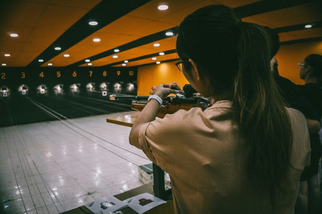 A person with long hair in a ponytail aims a rifle at numbered shooting targets in an indoor shooting range with yellow-orange ceiling lights. Two others stand nearby, partially visible—a perfect scene for any Shooting Range Directory.