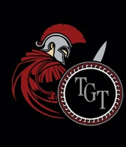 A stylized Roman or Greek soldier in red armor and helmet holds a round shield displaying the letters "TGT" on a black background.