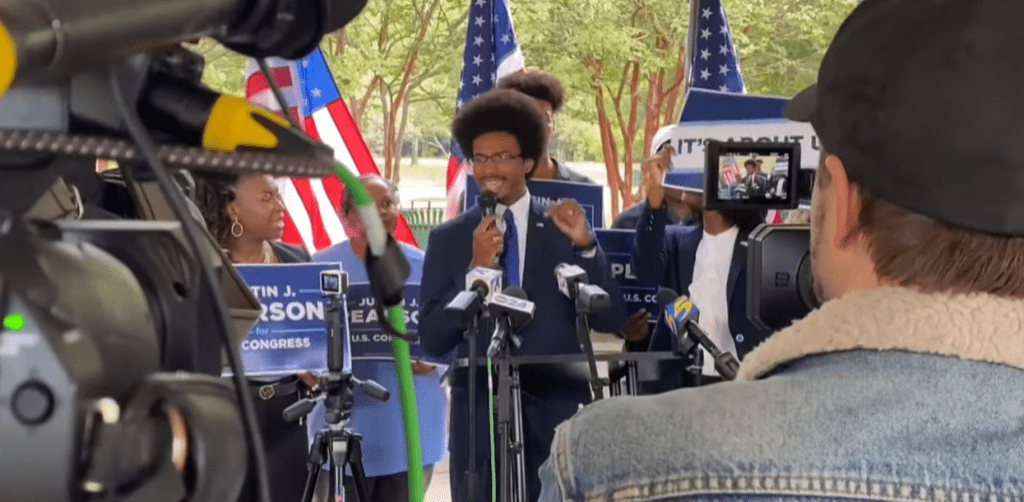 The Political Wave Changing Gun Safety Policies 1 A man in a suit speaks at a podium with microphones during an outdoor press conference, surrounded by supporters holding campaign signs and American flags, while cameras and a person in a cap film the event.