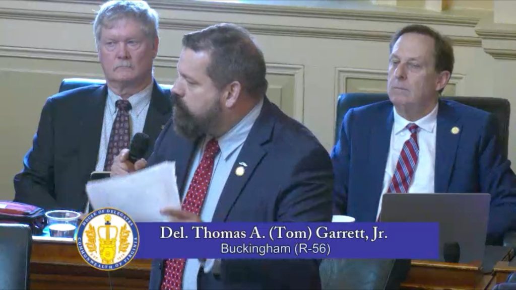 A man in a suit speaks into a microphone while holding papers, seated between two other men in suits. A caption reads, "Del. Thomas A. (Tom) Garrett, Jr. Buckingham (R-56)" with a seal on the left side.