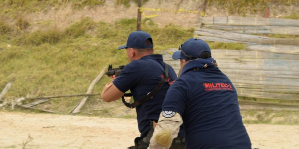 Two men in navy shirts and caps are outdoors on a shooting range. One is aiming a rifle ahead, while the other stands close behind, possibly instructing or observing. Both wear MILITEC-branded shirts and tactical gear.