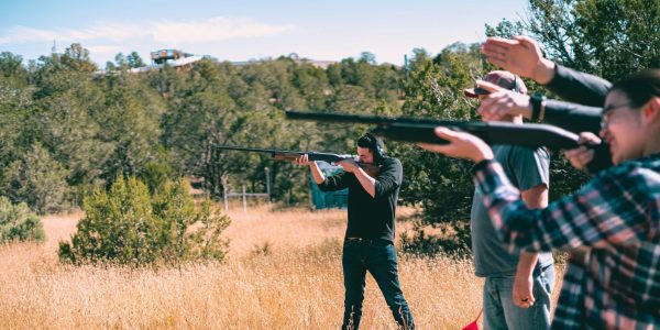 Four people stand in a grassy field aiming rifles towards the sky, surrounded by trees and under a clear blue sky. The group appears focused, with one person wearing ear protection.