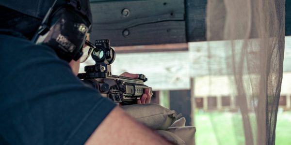A person wearing hearing protection aims a rifle with a scope through a window at an outdoor shooting range, resting the firearm on a sandbag for stability.
