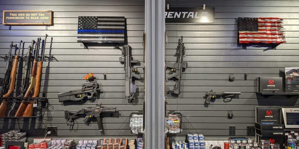 A display of firearms, ammunition, and accessories on black wall panels in a gun shop, with two American flag designs and a sign reading "Free men do not ask permission to bear arms.